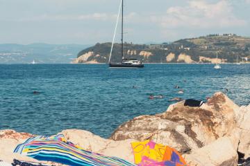 Beach towels on rocks with a sailboat in the distance on a bright day in Piran, Slovenia.