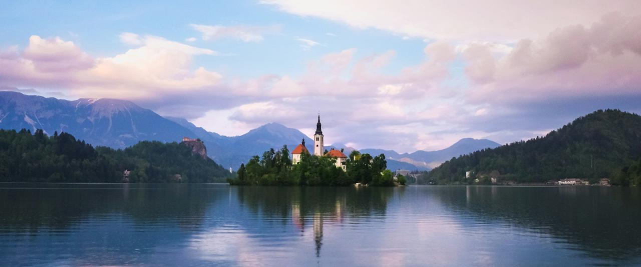 A scenic view of a lake with a small island featuring a church with a tall steeple, surrounded by mountains and reflecting in the water under a cloudy sky.