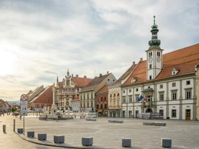 A wide, paved public square lined with historic European-style buildings featuring colorful facades and red-tiled roofs, dominated on the right by a prominent white building with a tall, ornate green-domed clock tower; a fountain is visible in the