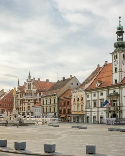 A wide, paved public square lined with historic European-style buildings featuring colorful facades and red-tiled roofs, dominated on the right by a prominent white building with a tall, ornate green-domed clock tower; a fountain is visible in the