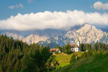 A white church with a green spire sits on a grassy hillside in a mountainous region, with a large forested slope in the foreground and a prominent rocky mountain range in the background.