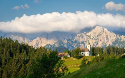 A white church with a green spire sits on a grassy hillside in a mountainous region, with a large forested slope in the foreground and a prominent rocky mountain range in the background.