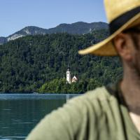 A man wearing a straw hat and glasses is in the foreground, with a view of a small island featuring a church with a tall steeple on a lake surrounded by forested mountains in the background.