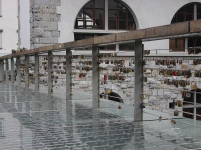 A modern pedestrian bridge with a glass floor and metal railings, covered in numerous padlocks (love locks), adjacent to a stone building with arched windows.
