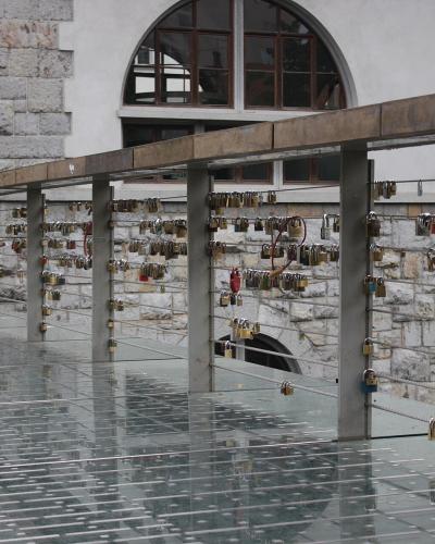 A modern pedestrian bridge with a glass floor and metal railings, covered in numerous padlocks (love locks), adjacent to a stone building with arched windows.
