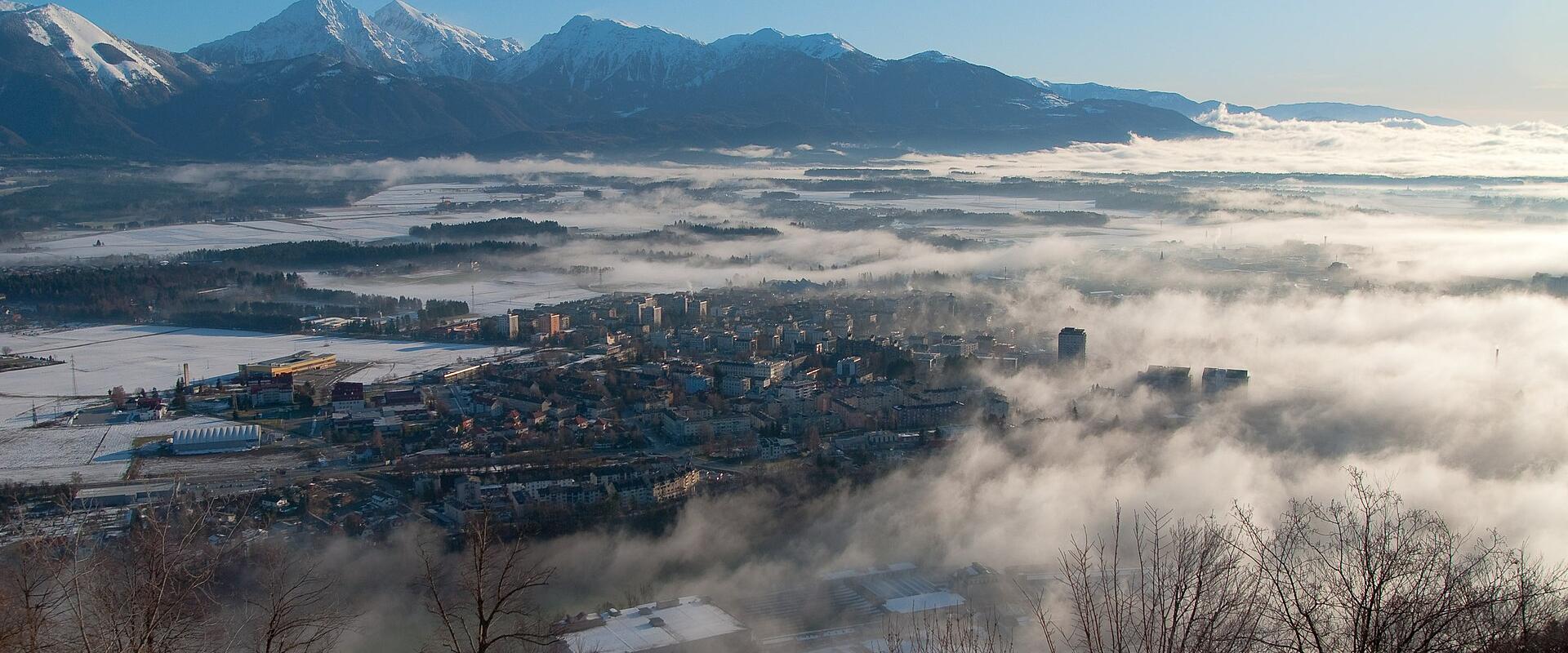 A panoramic view of Kranj, Slovenia, with the city nestled in a valley surrounded by snow-capped mountains and low-lying fog.