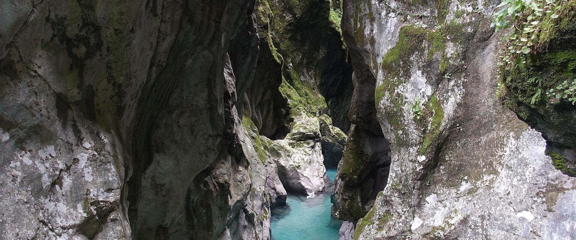 A narrow limestone gorge with steep, moss-covered rock walls enclosing a stream of clear, turquoise water flowing over a rocky bed.