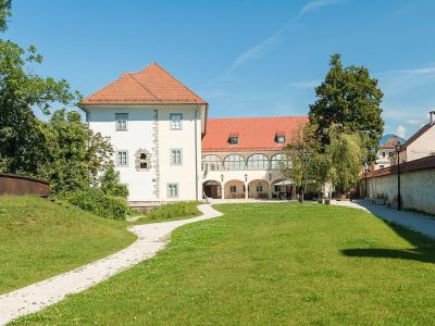 A white building with a red-tiled roof and arched loggia sits behind a grassy lawn, flanked by a stone wall and a modern rusted metal sculpture on the left.