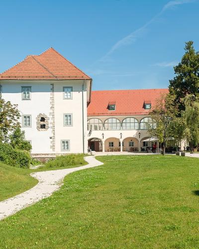 A white building with a red-tiled roof and arched loggia sits behind a grassy lawn, flanked by a stone wall and a modern rusted metal sculpture on the left.