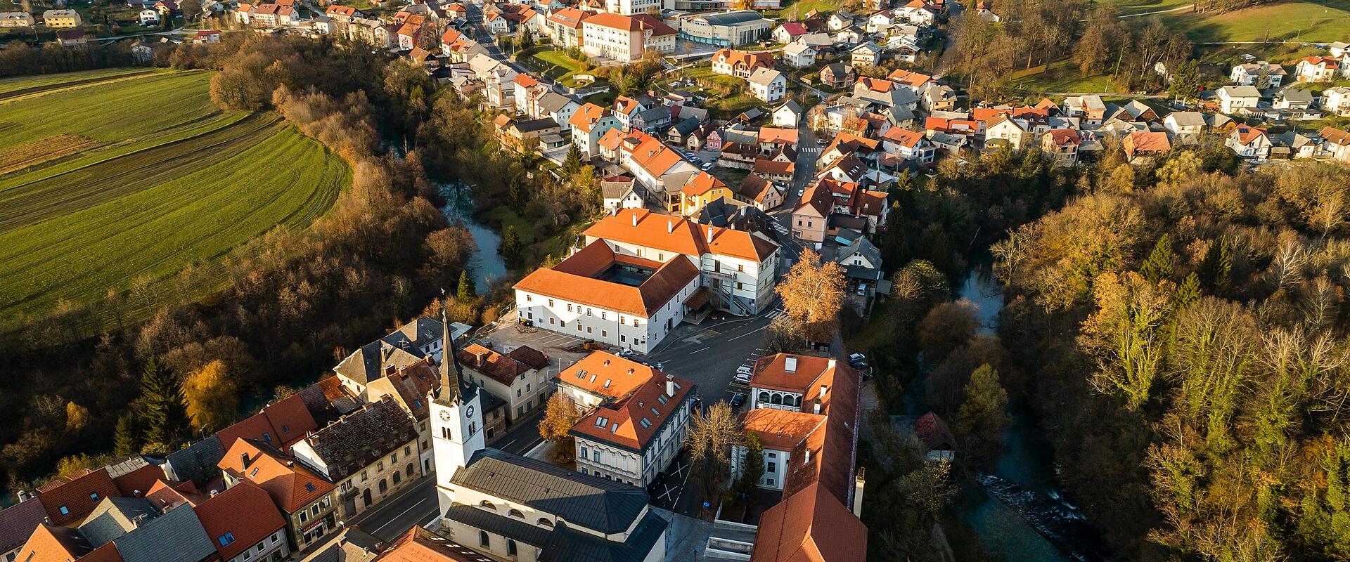 Aerial view of a town with red-roofed buildings, a prominent church, and a river, surrounded by green fields and trees.