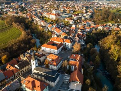 Aerial view of a town with red-roofed buildings, a prominent church, and a river, surrounded by green fields and trees.