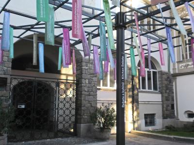The image shows the exterior facade of a white, multi-story historic building with arched loggia on the upper level, flower boxes, decorative quatrefoil windows, and two tall flagpoles flying Slovenian flags; vertical banners with partial text “PO