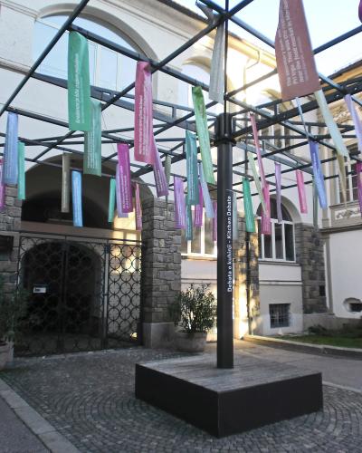 The image shows the exterior facade of a white, multi-story historic building with arched loggia on the upper level, flower boxes, decorative quatrefoil windows, and two tall flagpoles flying Slovenian flags; vertical banners with partial text “PO