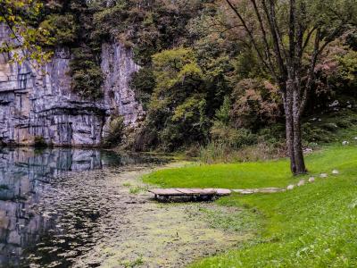 A scenic view of a calm river or spring with a steep, layered limestone cliff face on the left, a wooden platform extending into the water, and a grassy bank with benches and trees on the right.