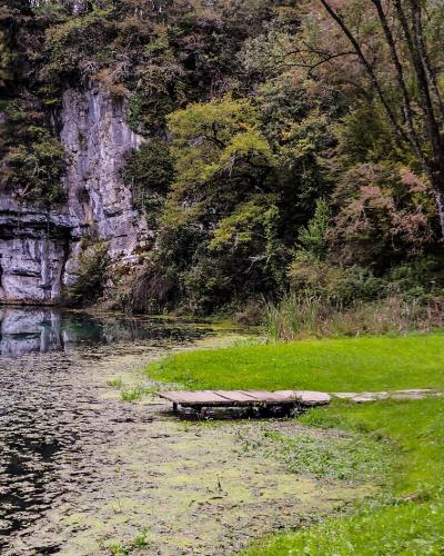 A scenic view of a calm river or spring with a steep, layered limestone cliff face on the left, a wooden platform extending into the water, and a grassy bank with benches and trees on the right.