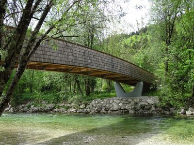 A long, wooden-shingled pedestrian and cyclist bridge spanning a clear, shallow river, supported by a distinctive V-shaped concrete pier, surrounded by dense green trees and a rocky riverbank.