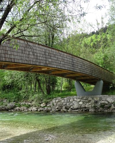 A long, wooden-shingled pedestrian and cyclist bridge spanning a clear, shallow river, supported by a distinctive V-shaped concrete pier, surrounded by dense green trees and a rocky riverbank.