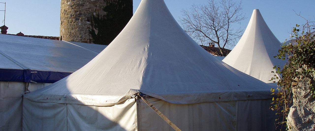 A stone medieval tower stands behind several large white event tents under a clear blue sky, with bare trees and a stone wall visible on the right.