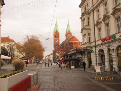 A pedestrian street scene in Maribor, Slovenia, featuring the distinctive red-brick Church of the Assumption of Mary with its twin green-spired towers in the background, a SPAR supermarket on the right, cobblestone paving, and a date stamp of 28/1