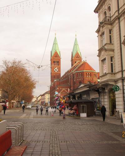 A pedestrian street scene in Maribor, Slovenia, featuring the distinctive red-brick Church of the Assumption of Mary with its twin green-spired towers in the background, a SPAR supermarket on the right, cobblestone paving, and a date stamp of 28/1