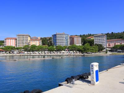 A daytime view of a marina or harbor with a concrete pier in the foreground, looking across the water at a row of large hotels and a beach area lined with trees and umbrellas.
