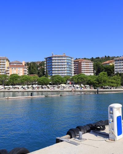 A daytime view of a marina or harbor with a concrete pier in the foreground, looking across the water at a row of large hotels and a beach area lined with trees and umbrellas.