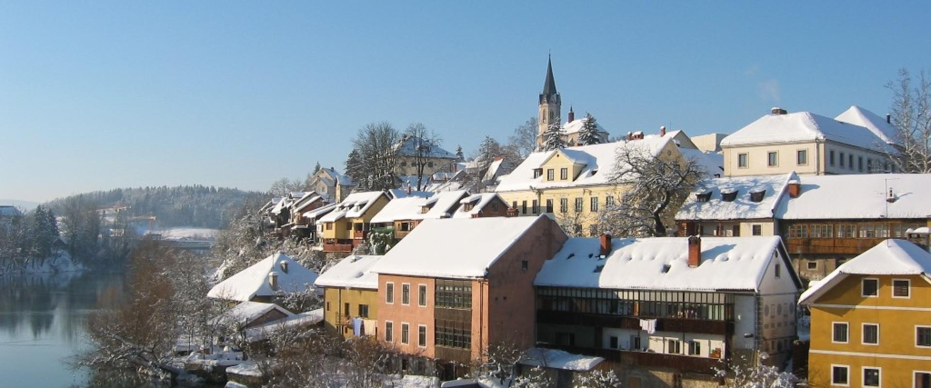A snow-covered riverside town scene featuring colorful buildings along a riverbank with a prominent church spire in the background.
