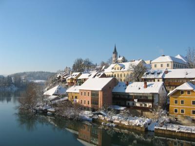 A snow-covered riverside town scene featuring colorful buildings along a riverbank with a prominent church spire in the background.