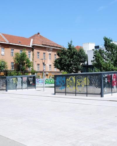 The image shows a paved public square with low, glass-paneled structures covered in graffiti, set against a backdrop of a large, historic pinkish-orange building with a red-tiled roof and modern white buildings, under a clear blue sky.