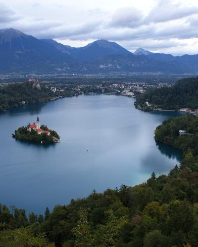 A high-angle panoramic photograph of Lake Bled featuring the island with the Church of the Assumption, Bled Castle on a cliff to the left, and the Julian Alps in the background.