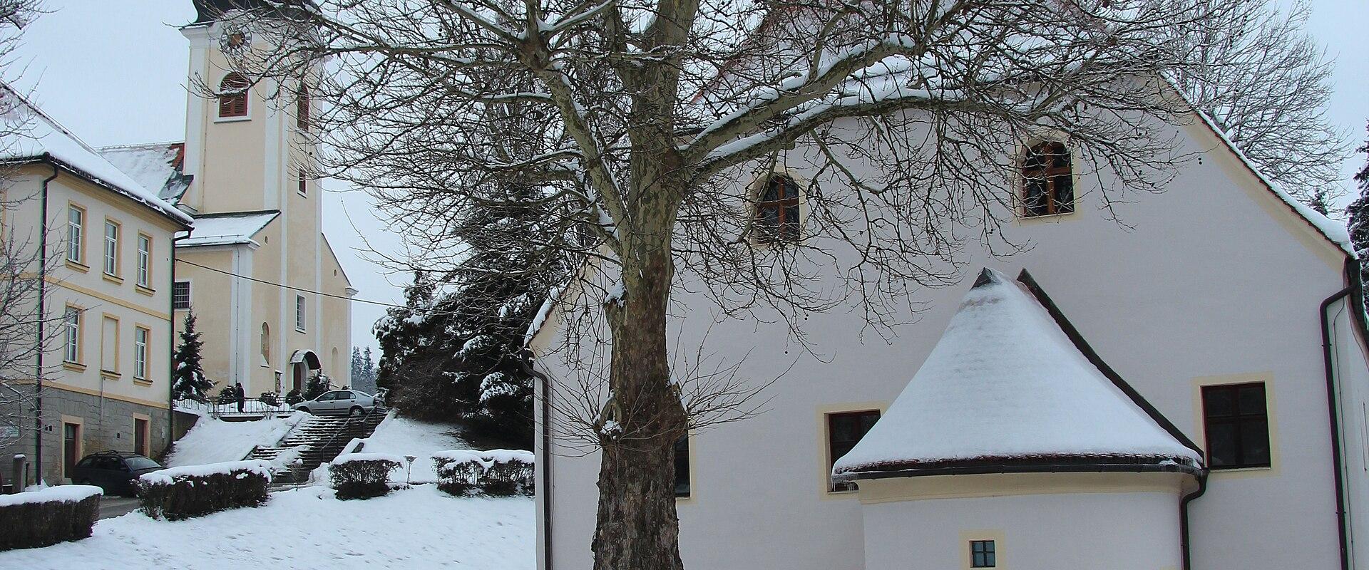 A snowy winter scene featuring a large bare tree in the foreground, a white building with a small conical roof extension to the right, and a church tower with a dark spire visible in the background on the left.