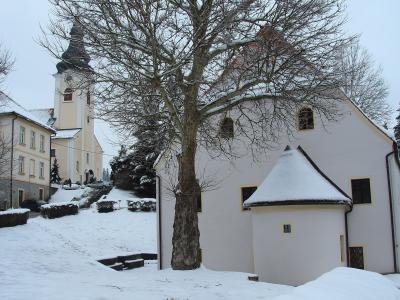 A snowy winter scene featuring a large bare tree in the foreground, a white building with a small conical roof extension to the right, and a church tower with a dark spire visible in the background on the left.