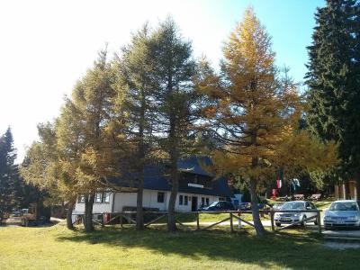 A white building with a dark roof partially obscured by tall trees with yellowing foliage, featuring a sign above the entrance that reads "RUŠKA KOČA" and several cars parked in front.