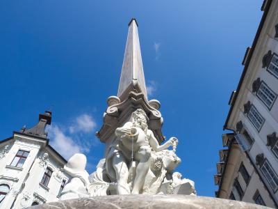 A low-angle shot of a Baroque stone fountain featuring a central obelisk and sculpted figures, set against a blue sky with adjacent historic buildings.