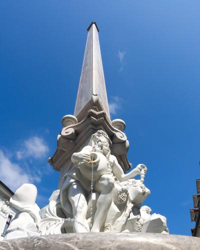 A low-angle shot of a Baroque stone fountain featuring a central obelisk and sculpted figures, set against a blue sky with adjacent historic buildings.