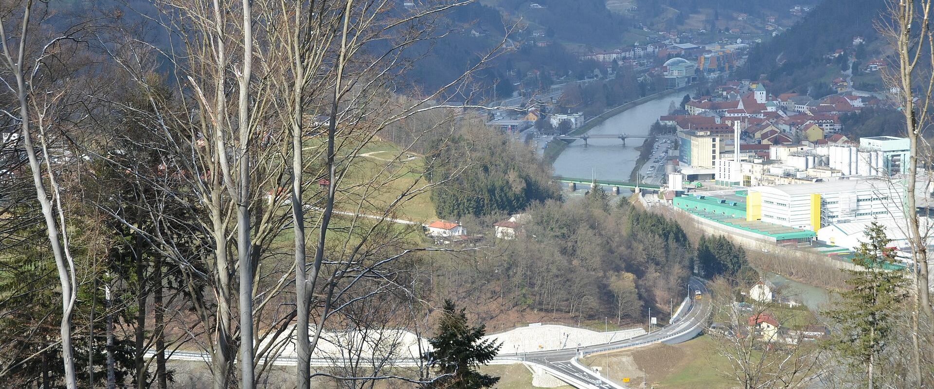 A high-angle landscape view showing a river valley with a bridge in the foreground, a town in the mid-ground, and a large industrial complex with a tall chimney on the right bank.