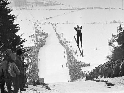 A black and white photograph of a ski jumper in mid-flight over a large, snow-covered ski jump hill, with massive crowds lining the landing area and spectators watching from the foreground.