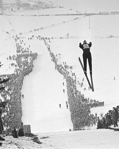 A black and white photograph of a ski jumper in mid-flight over a large, snow-covered ski jump hill, with massive crowds lining the landing area and spectators watching from the foreground.