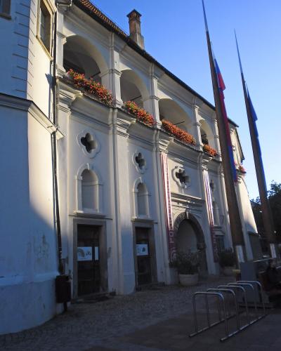 A view of a European town square with outdoor café seating in the foreground, featuring a prominent white building with a red-tiled roof and a distinctive dark spire, alongside other historic buildings and a plague column.