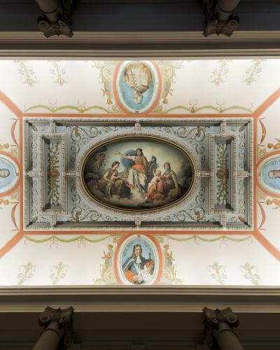 The image shows an ornate, painted ceiling with a central oval fresco depicting allegorical figures, surrounded by four oval portraits of historical men, framed by decorative stucco and painted garlands, viewed from below within a grand interior s