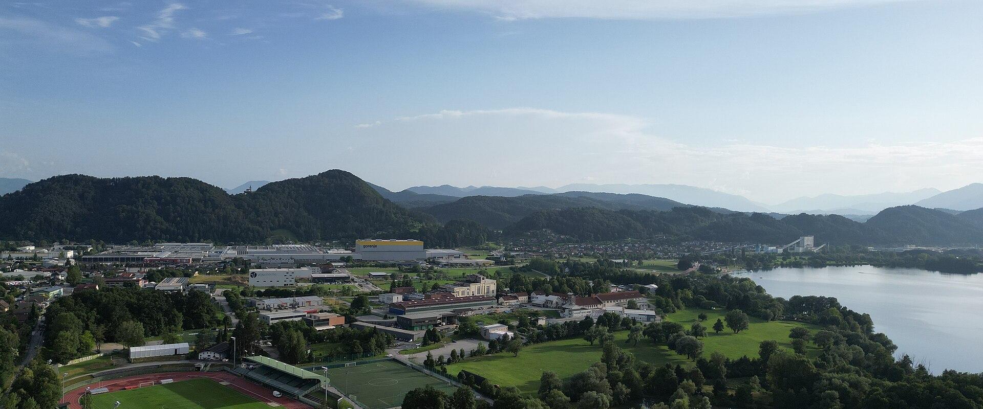 An aerial view of a town nestled in a valley next to a large body of water, featuring a prominent sports stadium with a red running track in the foreground and forested hills in the background.