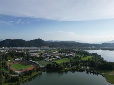 An aerial view of a town nestled in a valley next to a large body of water, featuring a prominent sports stadium with a red running track in the foreground and forested hills in the background.