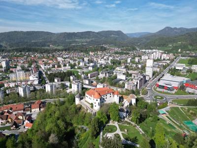 An aerial view of Velenje Castle situated on a green hill overlooking the city of Velenje, Slovenia, with residential buildings and mountains in the background.