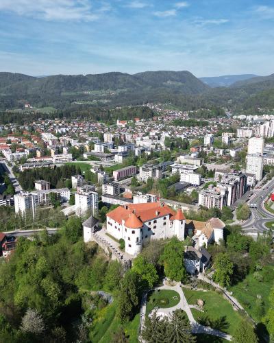 An aerial view of Velenje Castle situated on a green hill overlooking the city of Velenje, Slovenia, with residential buildings and mountains in the background.