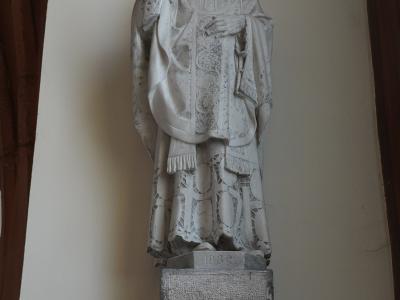 A white stone statue of a robed religious figure with a halo, mounted on a pedestal against a plain wall, with the date "1932" visible on the base.