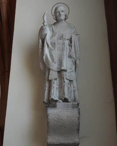A white stone statue of a robed religious figure with a halo, mounted on a pedestal against a plain wall, with the date "1932" visible on the base.