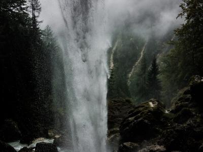 A tall, powerful waterfall plunging vertically into a rocky basin, surrounded by dark coniferous trees and heavy mist, with no visible signage or human structures.