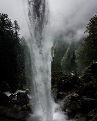 A tall, powerful waterfall plunging vertically into a rocky basin, surrounded by dark coniferous trees and heavy mist, with no visible signage or human structures.