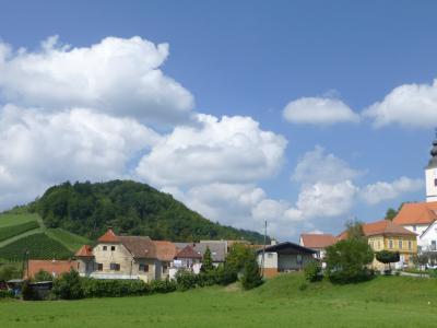 A rural village scene with a prominent white church tower, terraced vineyards on a green hillside, traditional houses with red-tiled roofs, and a bright blue sky with cumulus clouds; no visible signage or text identifying the location.