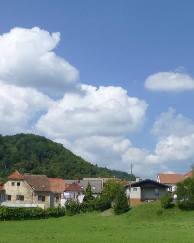 A rural village scene with a prominent white church tower, terraced vineyards on a green hillside, traditional houses with red-tiled roofs, and a bright blue sky with cumulus clouds; no visible signage or text identifying the location.